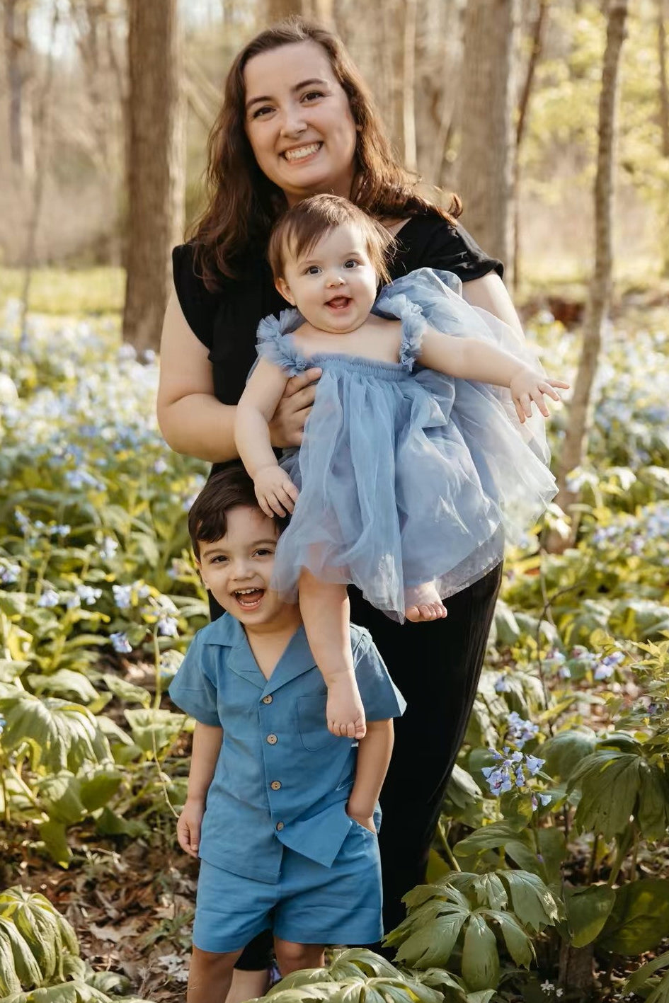 Woman holding a baby and standing next to a child in a forest with flowers
